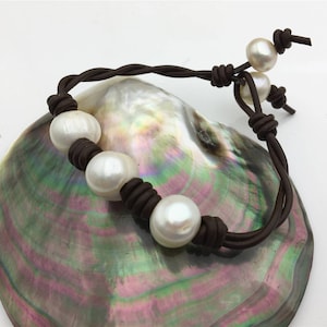 May include: A brown leather bracelet with five white pearl beads. The bracelet is displayed on a iridescent abalone shell.
