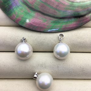 Pode incluir: Três pingentes de pérolas brancas com argolas prateadas são exibidos em um fundo neutro. As pérolas redondas são brilhantes e refletem a luz. Um grande fragmento de concha iridescente é visível ao fundo.