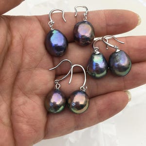 Puede incluir: Seis pares de pendientes de perlas iridiscentes en forma de lágrima con ganchos de plata. Las perlas muestran una gama de colores, incluyendo púrpura, azul y verde, con un brillo lustroso. Los pendientes se muestran sobre un fondo neutro.
