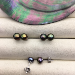 May include: Three pairs of dark iridescent pearl earrings. Two pairs are stud earrings with two pearls each. The third pair is a stud earring with two pearls and a post. The background is a light beige surface with a colorful iridescent shell.