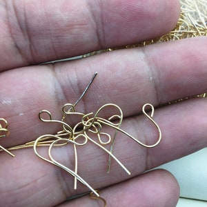 Puede incluir: Primer plano de un montón de ganchos para pendientes de color dorado. Los ganchos están hechos de metal fino y tienen un diseño simple y elegante.