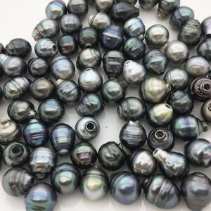 May include: A collection of dark green and black iridescent Tahitian pearls. The pearls are round and have a smooth, glossy finish. They are arranged in a loose pile on a white surface.