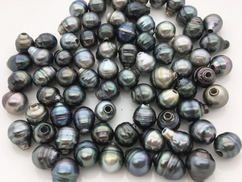 May include: A collection of dark green and black iridescent Tahitian pearls. The pearls are round and have a smooth, glossy finish. They are arranged in a loose pile on a white surface.