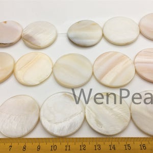 May include: A strand of white and cream colored round shell beads. The beads are approximately 1 inch in diameter and have a natural, iridescent sheen. The beads are strung on a white thread and are laid out on a ruler for size reference.