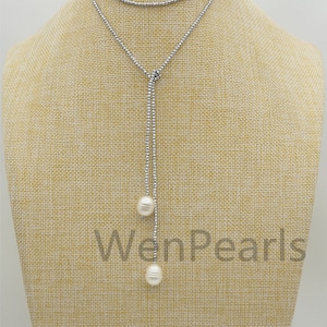 Op de afbeelding: Een zilveren ketting met twee witte parels die eronder hangen. De ketting is gelaagd en heeft een lang, hangend ontwerp. De ketting wordt getoond op een tan gekleurde stoffen mannequin.