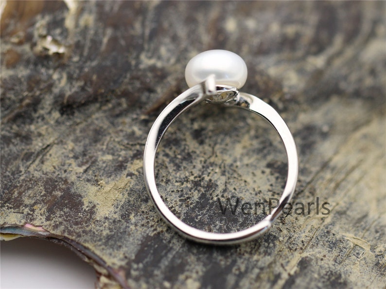 May include: A silver ring with a white pearl set on top. The ring is on a dark brown surface with the text "WenPearls" visible.