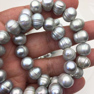 Puede incluir: Un collar de perlas grises grandes y brillantes. Las perlas tienen forma irregular, con una textura estriada y un brillo suave. Las perlas están ensartadas, creando un collar o una pulsera. Las perlas miden aproximadamente 10-12 mm de diámetro.
