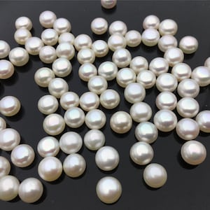 Puede incluir: Una colección de perlas redondas blancas esparcidas sobre una superficie negra. Las perlas son lisas y tienen un ligero brillo iridiscente.