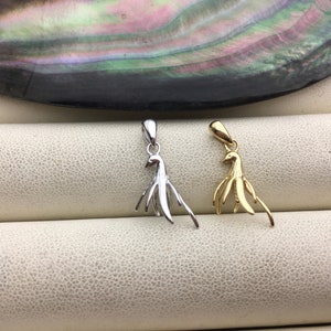 Puede incluir: Dos colgantes con forma de pájaro, uno plateado y otro dorado. Se presentan sobre una superficie texturizada de color blanco roto, con un fondo brillante e iridiscente. Los colgantes tienen un diseño estilizado de pájaro.