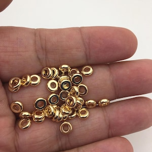 Puede incluir: Un conjunto de pequeñas cuentas metálicas doradas y brillantes. Las cuentas son redondas con un agujero en el centro, y están dispersas en una mano. Estas cuentas probablemente se utilizan para la fabricación de joyas u otros proyectos de artesanía.