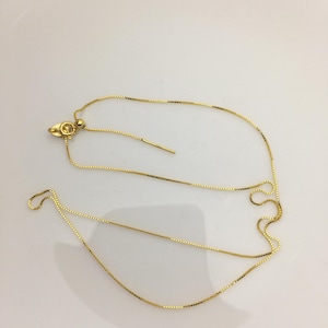 Può includere: Una delicata collana color oro con un design a catena a scatola. La collana presenta una lunghezza regolabile con una piccola sfera e una barra sottile. La chiusura è a forma di artiglio di aragosta. La catena è su una superficie bianca.