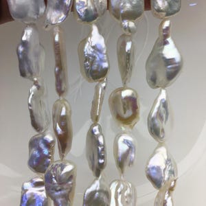 May include: A collection of white baroque pearls arranged in five vertical strands. Each pearl features an irregular, organic shape with a high-luster, iridescent surface that reflects subtle hints of pink, blue, and yellow light throughout.