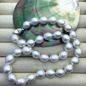 Puede incluir: Un collar de perlas blancas con un cierre plateado, exhibido sobre una concha nacarada e iridiscente. Las perlas son grandes y redondas, con un brillo lustroso. El fondo es una superficie neutra, blanquecina.