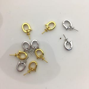 Pu&ograve; includere: Una collezione di chiusure per gioielli in tonalit&agrave; oro e argento. Le chiusure hanno un design unico con un anello e un piccolo perno per attaccare perline o altri componenti di gioielli. Le chiusure sono sparse su una superficie bianca.