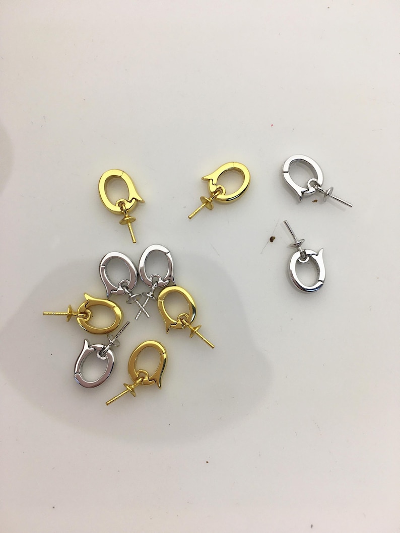 Pu&ograve; includere: Una collezione di chiusure per gioielli in tonalit&agrave; oro e argento. Le chiusure hanno un design unico con un anello e un piccolo perno per attaccare perline o altri componenti di gioielli. Le chiusure sono sparse su una superficie bianca.
