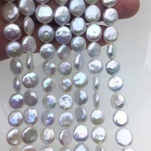 May include: Strands of lustrous, white, coin-shaped pearls. The pearls have a subtle iridescent sheen, reflecting hints of blue and purple. The pearls are strung together, ready for jewelry making.