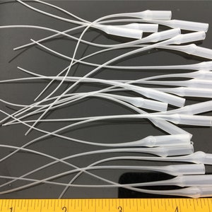 May include: A group of 20 clear plastic tubes with a tapered tip. The tubes are approximately 5 inches long and are arranged in a random pattern on a black surface. A ruler is visible in the bottom of the image.