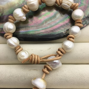 Könnte beinhalten: Ein handgefertigtes Armband mit großen, weißen Perlen, die auf einem hellbraunen Lederband aufgefädelt sind. Die Perlen sind mit dem Leder verknotet, wodurch ein einzigartiges Design entsteht. Das Armband wird auf einem neutralen Hintergrund präsentiert. Die Perlen messen ca. 1 cm im Durchmesser.