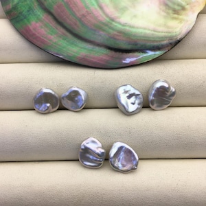 Puede incluir: Seis perlas iridiscentes de forma irregular, en tonos plateados y lavanda, se exhiben sobre una superficie color crema. Una gran concha iridiscente con tonos verdes, rosas y morados está en el fondo. Las perlas tienen una forma orgánica única.