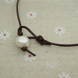 Puede incluir: Un collar sencillo de cuero marr&oacute;n con un solo colgante de perla blanca.