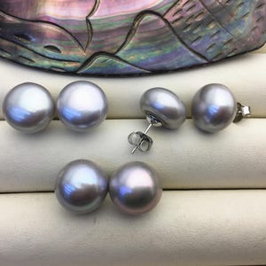 May include: Six lustrous, gray pearl earrings are displayed on a white surface. The earrings include two pairs of stud earrings and a pair of dangle earrings. The pearls have a smooth, iridescent surface, reflecting light with hints of blue and pink.