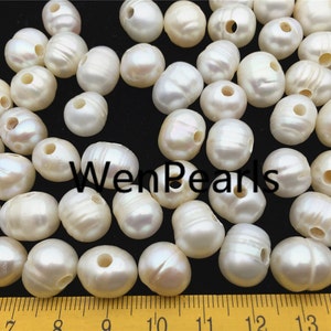May include: A collection of white, round, drilled pearls with a slightly irregular shape. The pearls are scattered on a black surface with a ruler in the foreground.