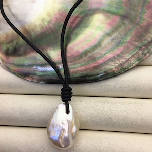 May include: A white baroque pearl pendant necklace with a black leather cord. The pearl has a lustrous surface and irregular shape. The cord is knotted at the top. The background features a mother-of-pearl shell.
