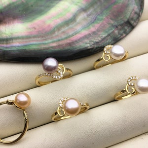 Może przedstawiać: Kilka złotych pierścionków z perłami i małymi kamieniami akcentującymi. Pierścionki mają delikatny design, z perłami w różnych kolorach, w tym białym, różowym i lawendowym. Pierścionki są prezentowane na białej powierzchni.