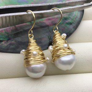 Puede incluir: Un par de pendientes envueltos en alambre dorado. Cada pendiente presenta una perla grande, blanca, en forma de lágrima, rodeada por alambre dorado enrollado y adornada con pequeñas perlas blancas. Los pendientes tienen cierres de gancho dorados.