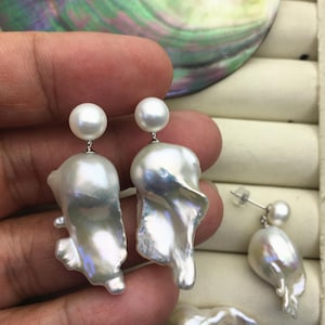 Könnte beinhalten: Weiße Perlenohrringe mit einem kleinen runden Perlenstecker und einem großen, unregelmäßig geformten Perltropfen. Die Ohrringe haben einen silberfarbenen Metallstift. Die Perlen haben einen schillernden Glanz.