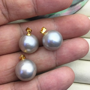 以下が含まれることがあります： 金色のバチカンが付いた、丸く光沢のあるグレーの真珠が3つ。真珠は滑らかで反射する表面を持っています。真珠は手で持たれており、そのサイズと色を示しています。金のバチカンが真珠にエレガントさを加えています。