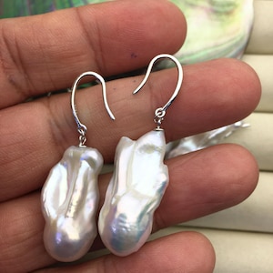 Può includere: Un paio di orecchini di perle barocche bianche con ganci color argento. Le perle hanno una forma irregolare e organica e una superficie lucida, che riflette la luce con sfumature iridescenti. Gli orecchini sono esposti su uno sfondo neutro.