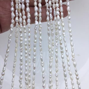 Puede incluir: Una hebra de perlas de agua dulce blancas, con una forma ligeramente irregular, enhebradas en un hilo fino. Las perlas están dispuestas en cascada, creando un aspecto delicado y elegante.