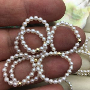Puede incluir: Primer plano de una mano sosteniendo varios hilos de collares de perlas blancas con detalles dorados. Los collares están hechos de pequeñas perlas redondas y están enhebrados con cuentas doradas.