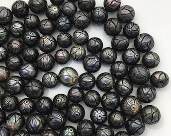 May include: A collection of carved black pearls with intricate floral designs. The pearls vary in size and have a lustrous, iridescent sheen, reflecting shades of grey, silver, and hints of purple. The pearls are arranged on a white surface.