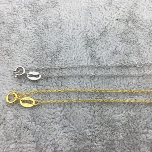以下が含まれることがあります： グレーのふわふわした表面に、銀と金の2本のチェーンが置かれています。両方のチェーンには、小さな楕円形のタグが付いた留め具があります。