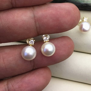 Könnte beinhalten: Ein Paar Perlenohrringe mit einem kleinen Diamanten-Akzent an jedem Ohrring. Die Ohrringe sind aus Gold und haben eine weiße Perle.