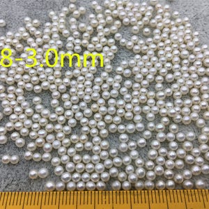 Może przedstawiać: Zbliżenie na stos białych, okrągłych pereł o średnicy 2,8-3,0 mm. Perły są rozrzucone na szarej powierzchni.