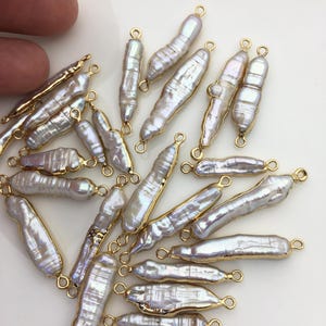 Könnte beinhalten: Eine Sammlung unregelmäßiger, länglicher weißer Perlen mit einem schimmernden Glanz. Jede Perle ist in einen goldfarbenen Metallrahmen mit einer kleinen Schlaufe an jedem Ende gefasst, was auf eine Verwendung in der Schmuckherstellung hindeutet. Die Perlen variieren in Größe und Form.