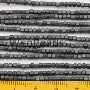 May include: A strand of dark gray square-shaped beads, approximately 4mm in size, with a ruler in the foreground for size reference.
