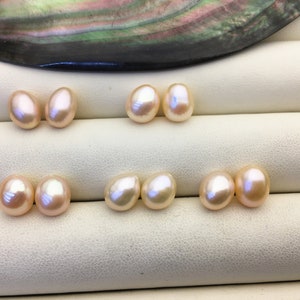 May include: Six oval-shaped pearls, arranged in three pairs, on a white surface. The pearls are a light pink color.