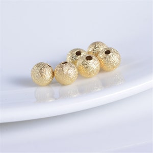 May include: A group of six gold-colored round beads with a textured surface. The beads have a hole in the center for stringing.