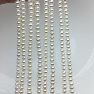 May include: Multiple strands of white pearls are shown, each with pearls of different sizes. The pearls are strung together, creating a flowing appearance. The pearls have a shiny surface, reflecting light. The background is a plain white surface.