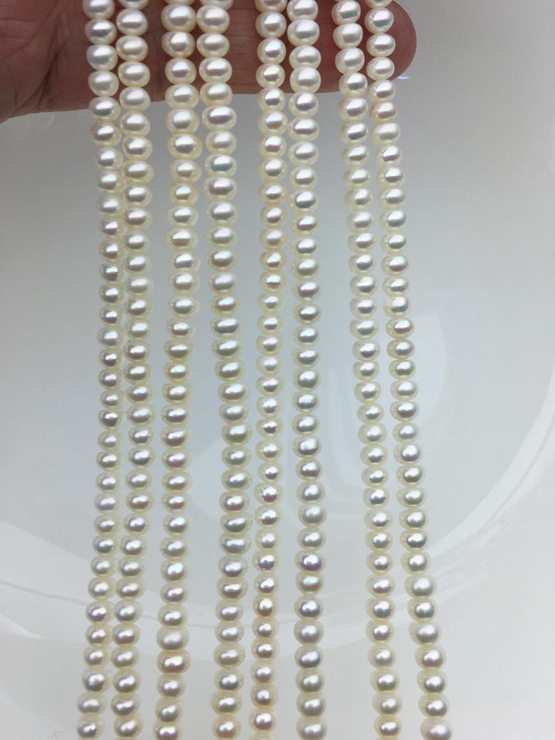 May include: Multiple strands of white pearls are shown, each with pearls of different sizes. The pearls are strung together, creating a flowing appearance. The pearls have a shiny surface, reflecting light. The background is a plain white surface.