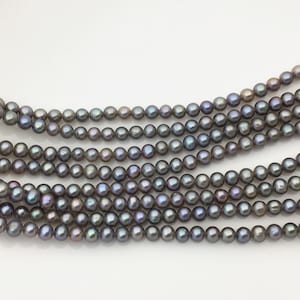 Puede incluir: Un hilo de pequeñas perlas redondas de color gris oscuro con matices iridiscentes en tonos morados y azules. Las perlas están ensartadas en una línea continua, mostrando una superficie lisa y brillante con un tamaño uniforme en todo el hilo.