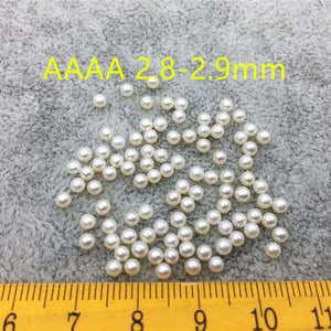 Op de afbeelding: Een close-up van een stapel witte ronde parels, met een diameter van 2,8-2,9 mm, op een grijs, pluche oppervlak. Een liniaal is zichtbaar aan de onderkant van de afbeelding, met markeringen van 5 tot 12.