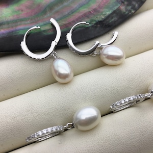 Könnte beinhalten: Ein Paar silberne Creolen mit jeweils einem weißen Perlenanhänger. Die Creolen sind mit kleinen, klaren Steinen verziert.