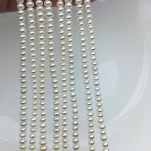 May include: Multiple strands of small, round, white pearls. The pearls are strung together, forming vertical lines. The pearls have a lustrous sheen, reflecting light. The background is a plain white surface, accentuating the pearls' colour and shape. These pearls are likely for jewellery making.