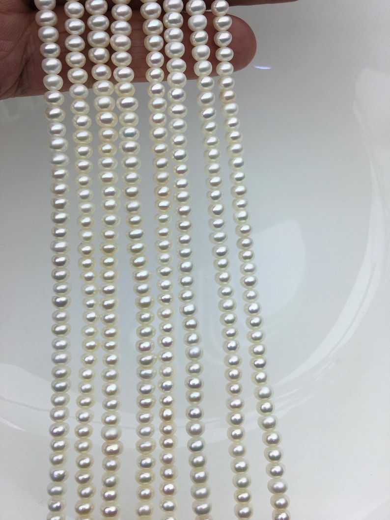 May include: Multiple strands of small, round, white pearls. The pearls are strung together, forming vertical lines. The pearls have a lustrous sheen, reflecting light. The background is a plain white surface, accentuating the pearls' colour and shape. These pearls are likely for jewellery making.