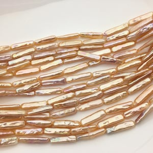 May include: Close-up of strands of rectangular, iridescent beads in shades of peach, gold, and lavender. The beads have a natural, organic shape and are arranged in parallel rows, creating a textured surface. The background is a soft, off-white color.